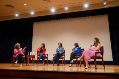 Five women sit in chairs and hold microphones on a small stage. Four of them look to the left at their fellow panel member, who wears a pink blouse and speaks into her microphone.